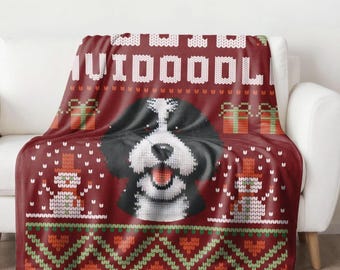 May include: A red blanket with a Bernedoodle dog design. The blanket features a knitted pattern with the word "Bernedoodle" in white, along with gift boxes and snowmen. The blanket is draped over a white sofa.