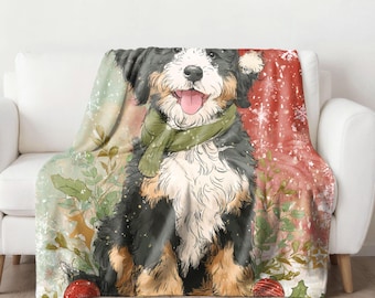 May include: A cozy throw blanket featuring a Bernese Mountain Dog illustration. The dog is wearing a green scarf and is surrounded by festive holiday elements, including red ornaments and snowflakes. The blanket is draped over a white sofa.
