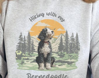 Puede incluir: Sudadera gris claro con un gráfico de un perro Bernedoodle en un paisaje montañoso. El texto dice "Hiking with my Bernedoodle". El perro es negro, blanco y gris con un collar rojo.