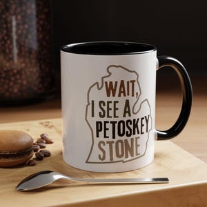 May include: White coffee mug with a black rim and handle. The mug features a brown outline of the state of Michigan with the text "WAIT, I SEE A PETOSKEY STONE" in brown lettering.