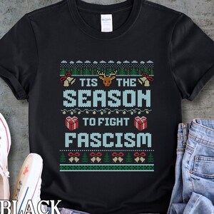 May include: Black t-shirt with a festive design. The text reads "TIS THE SEASON TO FIGHT FASCISM" in light blue, with Christmas-themed graphics. The shirt has a crew neck and short sleeves.