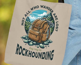 May include: Beige tote bag with black handles featuring a circular graphic of a brown backpack, mountains, and the text "NOT ALL WHO WANDER ARE LOST" and "ROCKHOUNDING". The bag is made of a durable material.