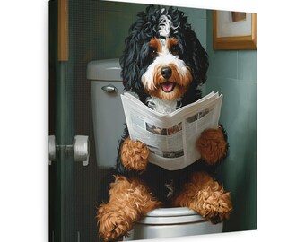 May include: A canvas print depicts a Bernedoodle dog sitting on a toilet, engrossed in reading a newspaper. The dog has black, white, and brown fur. The bathroom setting features a white toilet, a roll of toilet paper, and a framed picture on the wall.
