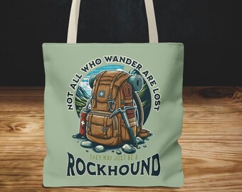 May include: A light green tote bag with a graphic of a brown backpack, ice axe, and other hiking gear. The text "NOT ALL WHO WANDER ARE LOST" and "ROCKHOUND" are printed on the bag. The bag has cream-colored handles.