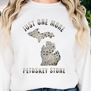 May include: White sweatshirt featuring a Petoskey stone design in the shape of the state of Michigan. The text "JUST ONE MORE" arches above the stone, with "PETOSKEY STONE" below. The stone design is a mix of gray and brown.