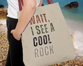 May include: Beige tote bag with the text "WAIT, I SEE A COOL ROCK" in various colors. The bag has red straps and is held by a person. The background shows a beach with rocks and the ocean.