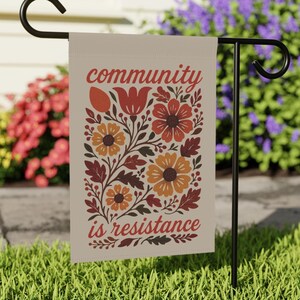 May include: A beige garden flag displaying the words "community is resistance" in red script. The flag features a floral design with orange, red, and brown flowers and leaves. The flag is hanging on a black metal stand in a garden.
