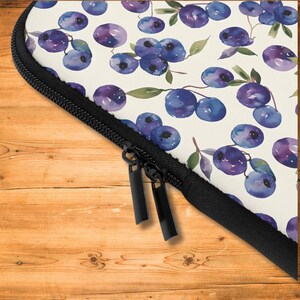 May include: A white laptop sleeve with a zipper closure. The sleeve features a watercolor pattern of purple blueberries and green leaves.