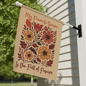 May include: Tan outdoor flag with floral design in shades of orange, red, and brown. The flag reads "My Favorite Season Is the Fall of Fascism" in a decorative font. The flag is mounted on a white pole.