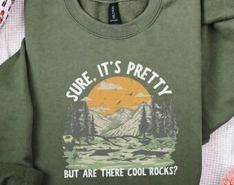 May include: Olive green sweatshirt with a graphic of a mountain landscape, a setting sun, and the text "Sure, it's pretty, but are there cool rocks?" The design features trees, mountains, and birds.