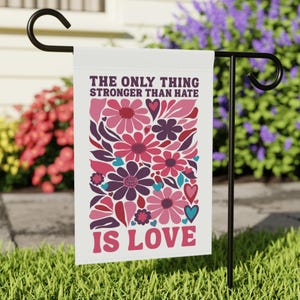 May include: A white garden flag with the text "THE ONLY THING STRONGER THAN HATE IS LOVE" in a retro font. The flag features a floral design in shades of pink, purple, and teal. The flag is displayed on a black metal stand in a garden.