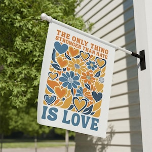 May include: A white flag with the text "THE ONLY THING STRONGER THAN HATE IS LOVE" in orange and blue. The flag features a floral design with orange and blue hearts and flowers. The flag is hanging outside.