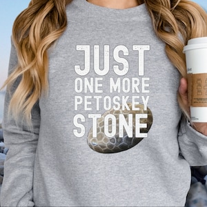 May include: A light gray sweatshirt with the text "JUST ONE MORE PETOSKEY STONE" in white block letters. A detailed image of a Petoskey stone is below the text. The sweatshirt is a casual style.