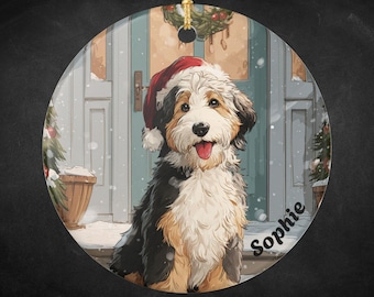 May include: Round Christmas ornament featuring a dog wearing a Santa hat. The dog has black, white, and brown fur and is sitting on a snowy porch. The name "Sophie" is written on the ornament.
