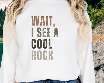 May include: White sweatshirt with the text "WAIT, I SEE A COOL ROCK" in brown, green, and gray lettering. The sweatshirt is paired with blue jeans. The background features a natural outdoor scene with trees and a body of water.