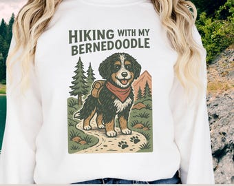 Puede incluir: Sudadera blanca con un gráfico de un perro Bernedoodle con mochila y pañuelo rojo, con el texto "Hiking with my Bernedoodle". El perro está en un camino con árboles y una montaña al fondo. La marca "Bananadoo Designs" está en la parte inferior.