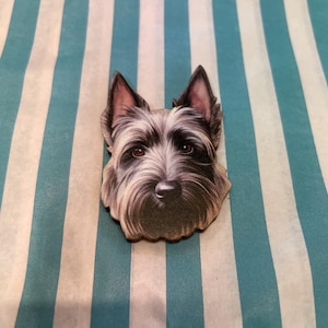 Könnte beinhalten: Ein Holzausschnitt eines Hundegesichts, der einen Terrier zeigt. Der Hund hat ein detailliertes schwarzes und graues Fell, braune Augen und eine lange Schnauze. Der Hintergrund ist ein gestreiftes Muster aus Weiß und Hellblau.