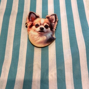Könnte beinhalten: Ein holzfarbener Chihuahua-Hunde-Ansteckpin mit einer detaillierten, farbenfrohen Illustration. Der Pin zeigt das Gesicht des Hundes mit braunem, weißem und hellbraunem Fell. Der Pin befindet sich auf einem gestreiften blau-weißen Hintergrund.