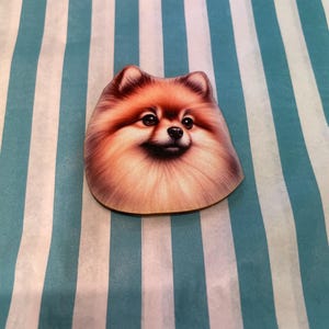 Könnte beinhalten: Holzausschnitt in Form eines Zwergspitzes mit orange-braunem Fell-Design. Der Hund hat einen lächelnden Gesichtsausdruck. Der Hintergrund ist blau und weiß gestreift.