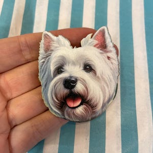 Könnte beinhalten: Anstecknadel mit dem Gesicht eines West Highland White Terrier-Hundes mit detailliertem, realistischem Design. Die Nadel zeigt das weiße Fell des Hundes, rosa Ohren und eine rosa Zunge. Das Gesicht des Hundes ist oval.