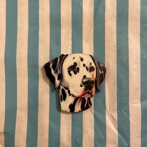 Könnte beinhalten: Ein dekoratives Objekt in Form eines Dalmatiner-Hundekopfes mit schwarzen Flecken auf weißem Grund. Der Hund befindet sich vor einem Hintergrund aus blau-weißen vertikalen Streifen.