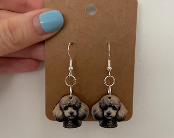 Puede incluir: Un par de pendientes con forma de caniche. Los pendientes tienen ganchos plateados y pequeños anillos plateados redondos. La cabeza del caniche está representada con una nariz y ojos negros, y un pelaje esponjoso, marrón claro y negro. Los pendientes están sujetos a un respaldo de cartón marrón.