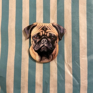 Könnte beinhalten: Ein holzförmiger Ansteckpin in Form einer Möpse mit einer detaillierten Illustration des Gesichts eines Mopses. Der Pin hat ein braunes und schwarzes Farbschema mit einem leicht mürrischen Ausdruck. Der Hintergrund ist ein blau-weiß gestreiftes Muster.