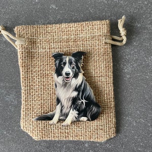Könnte beinhalten: Ein schwarz-weißer Border Collie, der auf einem Holzausschnitt gemalt ist. Der Ausschnitt liegt in einem kleinen Jutesack mit Kordel.