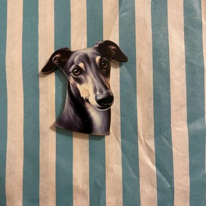 Könnte beinhalten: Ein Magnet in Form eines Greyhound-Hundes mit einem Schwarz-Grau-Farbschema. Der Kopf des Hundes ist der Mittelpunkt, mit detaillierten Augen und einer realistischen Darstellung. Der Magnet befindet sich vor einem Hintergrund aus vertikalen blau-weißen Streifen.