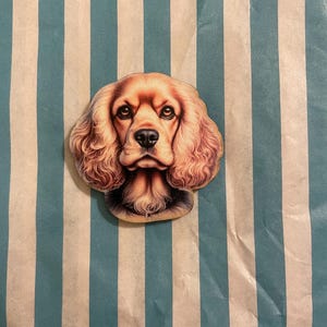 Könnte beinhalten: Ein Holzausschnitt eines Cocker Spaniel-Hundes mit detailliertem Fell und Gesichtszügen. Der Hund ist hellbraun mit dunkleren braunen Augen und einer schwarzen Nase. Der Ausschnitt befindet sich auf einem gestreiften Hintergrund in Türkis und Weiß.