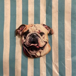 Könnte beinhalten: Ein Bulldoggen-förmiger Ansteckpin mit einem lächelnden Gesichtsausdruck. Der Pin zeigt eine detaillierte Illustration des Gesichts des Hundes, die seine Falten und Zunge zeigt. Der Hintergrund ist ein blau-weiß gestreiftes Muster.