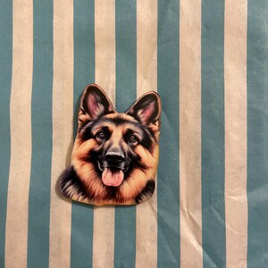 Könnte beinhalten: Ein Aufkleber in Form eines Schäferhundekopfes mit detailliertem, realistischem Design. Der Aufkleber zeigt das Gesicht des Hundes mit braunem, schwarzem und tanfarbenem Fell, rosa Zunge und aufmerksamem Ausdruck. Der Hintergrund ist ein blau-weiß gestreiftes Muster.