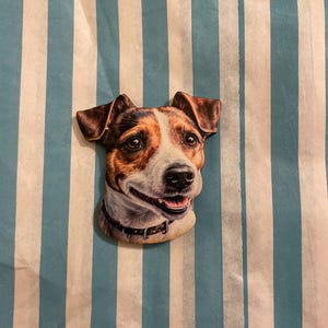 Könnte beinhalten: Ein Holzausschnitt eines Jack Russell Terriers mit braunem, schwarzem und weißem Fell. Der Hund lächelt mit leicht herausgestreckter Zunge. Der Ausschnitt befindet sich auf einem blau-weiß gestreiften Hintergrund.