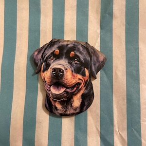 Könnte beinhalten: Aufkleber in Form eines lächelnden Rottweiler-Hundekopfes. Der Aufkleber ist schwarz und braun, wobei die Augen und der Mund des Hundes deutlich zu sehen sind. Der Hintergrund ist ein gestreiftes Muster aus Weiß und Hellblau.