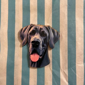 Könnte beinhalten: Ein Holzausschnitt des Kopfes einer Deutschen Dogge, mit einer detaillierten Illustration mit schwarzem Fell und rosa Zunge. Der Hund ist vor einem Hintergrund aus vertikalen Streifen in Hellblau und Weiß platziert.