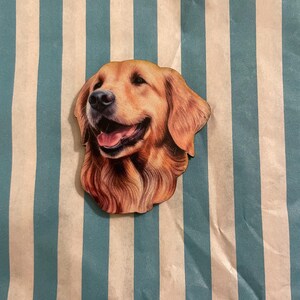Könnte beinhalten: Holzausschnitt eines Golden Retriever-Hundekopfes mit detailliertem Fell und fröhlichem Ausdruck. Der Hund ist hellbraun mit einer rosa Zunge. Der Hintergrund ist ein blau-weiß gestreiftes Muster.