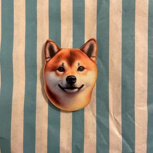 Könnte beinhalten: Aufkleber mit dem lächelnden Gesicht eines Shiba Inu-Hundes mit braun-weißem Fell. Der Hintergrund ist blau und weiß gestreift.