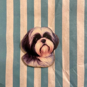 Könnte beinhalten: Holzausschnitt in Form eines Shih Tzu-Hundekopfes mit detaillierter Illustration. Der Hund hat schwarz-weißes Fell mit rosa Akzenten. Der Ausschnitt befindet sich auf einem blau-weiß gestreiften Hintergrund.