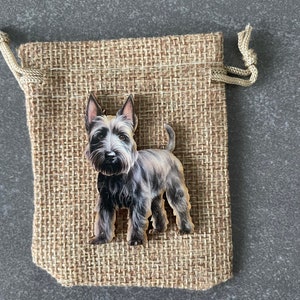 Könnte beinhalten: Ein hölzernes Hundeornament mit schwarzem und grauem Fell, platziert auf einem kleinen Jutebeutel. Der Beutel besteht aus gewebtem, beigem Material und hat einen Kordelzugverschluss. Der Hund hat ein detailliertes, realistisches Aussehen.