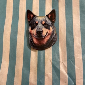 Könnte beinhalten: Ein Holzausschnitt eines Australian Cattle Dog mit einem lächelnden Gesichtsausdruck. Der Hund hat eine schwarze Schnauze, Ohren und Augen mit einer Mischung aus grauem, schwarzem und braunem Fell. Der Hintergrund ist ein gestreiftes Muster aus Türkis und Weiß.