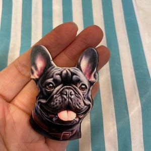 Könnte beinhalten: Ein schwarzer French-Bulldoggen-Kopf-Pin mit braunem Halsband und rosa Zunge. Der Pin wird in einer Hand vor einem Hintergrund aus blau-weiß gestreiftem Papier gehalten.