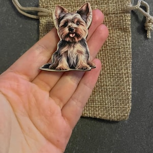 Könnte beinhalten: Ein hölzerner Magnet in Form eines Yorkie-Hundes mit detailliertem Fell und herausgestreckter Zunge. Der Magnet wird in einer Hand gehalten, neben einem kleinen Jute-Kordelzugbeutel. Der Hund hat braunes, schwarzes und hellbraunes Fell.