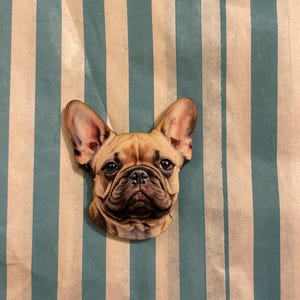Könnte beinhalten: Holzausschnitt eines französischen Bulldoggenkopfes mit detailliertem, realistischem Design. Der Hund hat ein hellbraunes Fell, dunkle Augen und ein faltiges Gesicht. Der Ausschnitt befindet sich vor einem gestreiften Hintergrund in Blau und Weiß.
