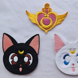May include: Embroidered patches featuring a gold and pink heart with wings, a black cat face with a yellow crescent moon, and a white cat face with blue eyes and a yellow crescent moon. The patches are on a white background.