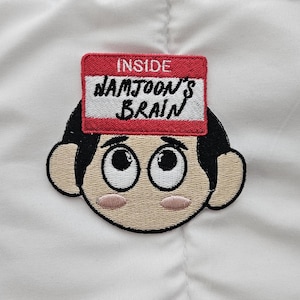May include: Embroidered patch featuring a cartoon face with large eyes and black hair. A red name tag above the head reads "INSIDE NAMJOON'S BRAIN" in white lettering. The patch is set against a white fabric background.