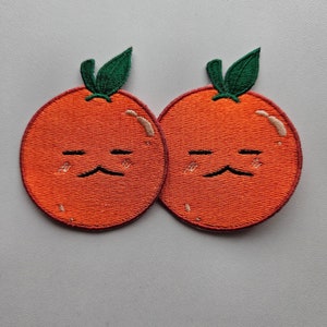 May include: Two orange embroidered patches with a cartoon orange design. Each patch features a sad face with a black mustache and two black lines for eyes. Green leaves are at the top of each orange.