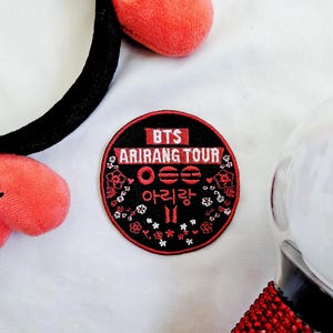 May include: A black headband with two red heart-shaped plushies, a round black and red patch with the text "BTS ARIRANG TOUR" in white and Korean characters, and a red and clear light stick.