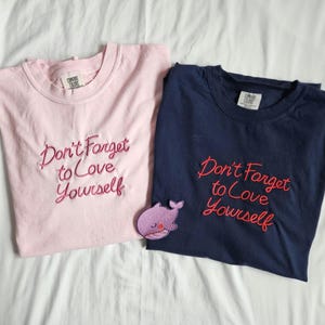 May include: Two t-shirts, one pink and one navy blue, with the embroidered phrase "Don't Forget to Love Yourself." A small purple whale patch is next to the navy blue shirt. Both shirts have a crew neck.