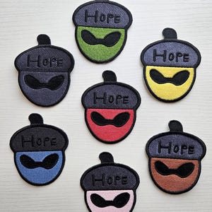 May include: Embroidered acorn patches in various colors, including green, yellow, red, blue, pink, and brown. Each patch features the word "HOPE" and a stylized face design. The patches are suitable for clothing or accessories.