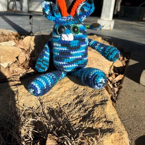 May include: A blue and black striped crocheted stuffed animal with orange horns and large green eyes. The animal is sitting on a rock.